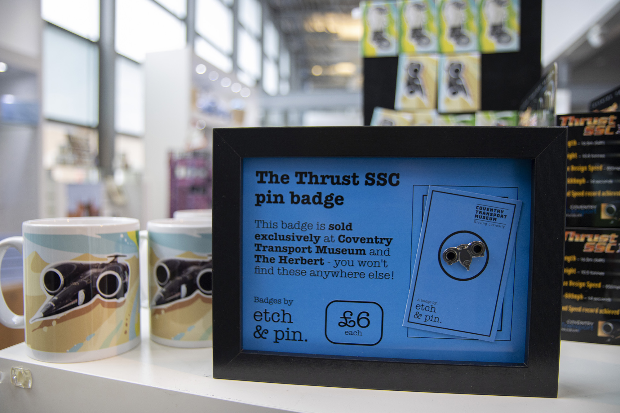 A photograph of Thrust SSC merchandise on display in the Coventry Transport Museum shop, including printed mugs, an Etch and Pin badge in a display frame, and boxes of model cars