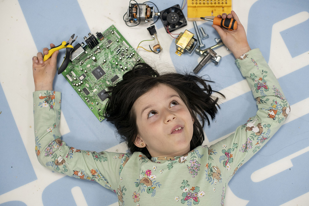 A smiling girl lying on the floor with pliers in one hand a screwdriver in the other. Surrounding her is a selection of nuts, bolts and electronic parts 