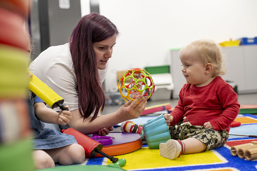 A woman holding a plastic ball with a tube of coloured beads inside it out towards a baby, who is sitting on the floor surrounded by other play items.