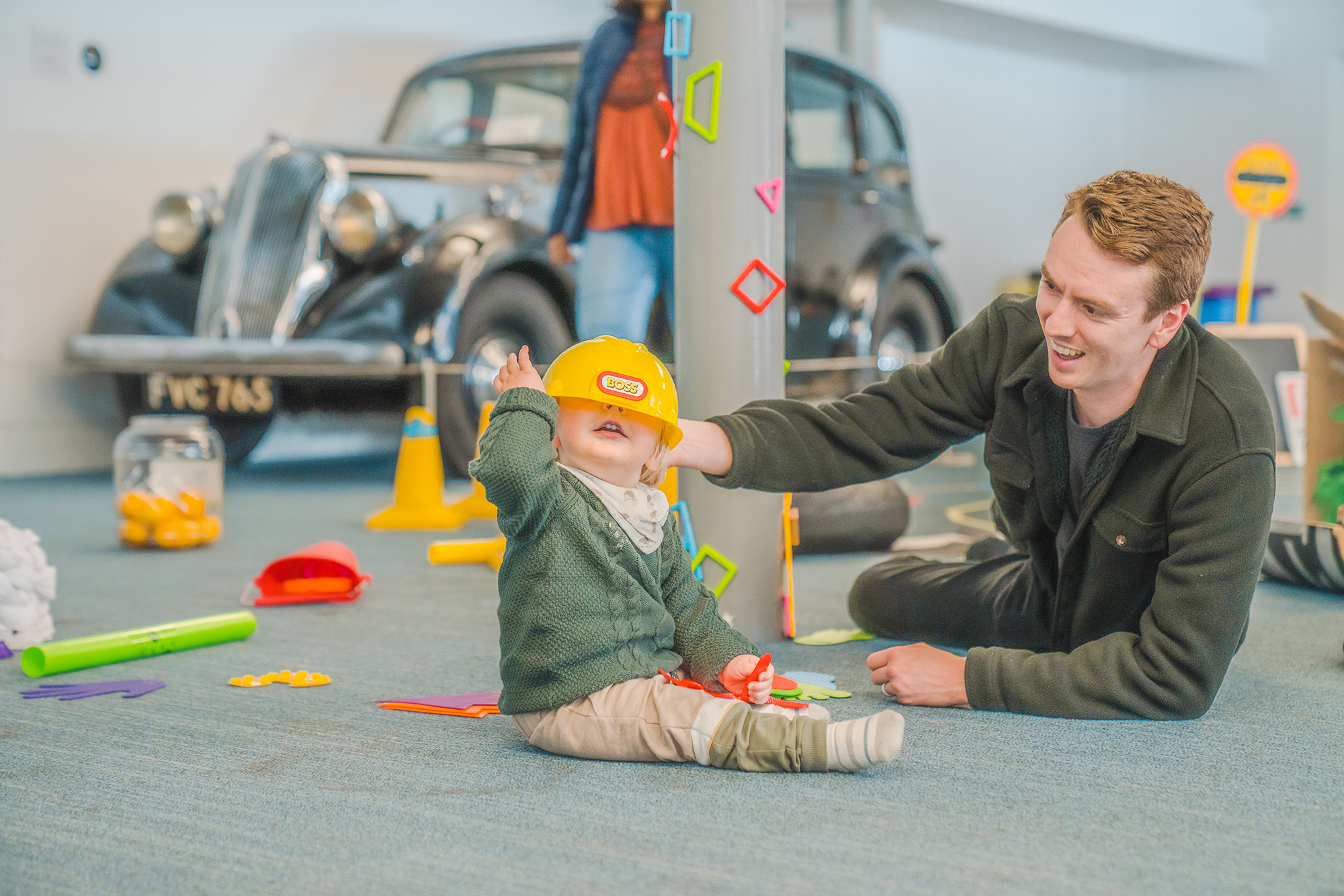 A man and a toddler playing with objects in front of a car on display at Coventry Transport Museum. The toddler has a yellow toy hard hat on his head, covering his eyes. There are toy traffic cones, magnets and a stop sign around them.