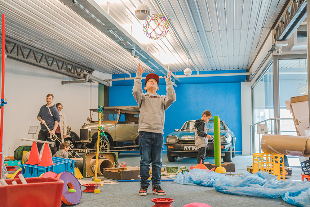 A boy raising his arms to catch a collapsible ball he has thrown up in the air. Around him are assorted play items and families using them.