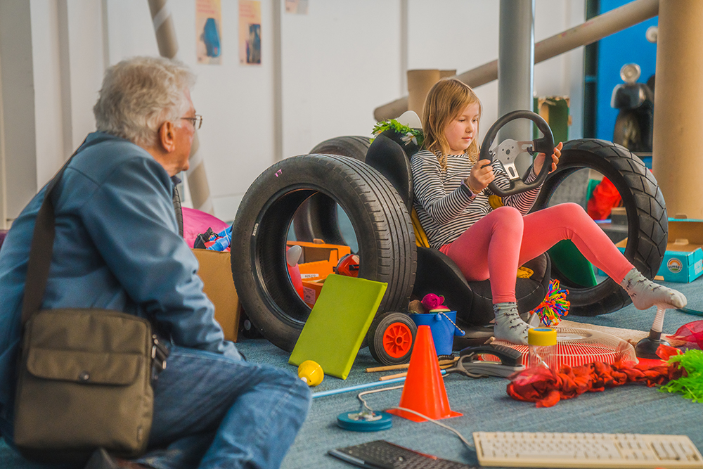 A girl sitting in a seat surrounded by tyres, holding a steering wheel while her grandad watches.