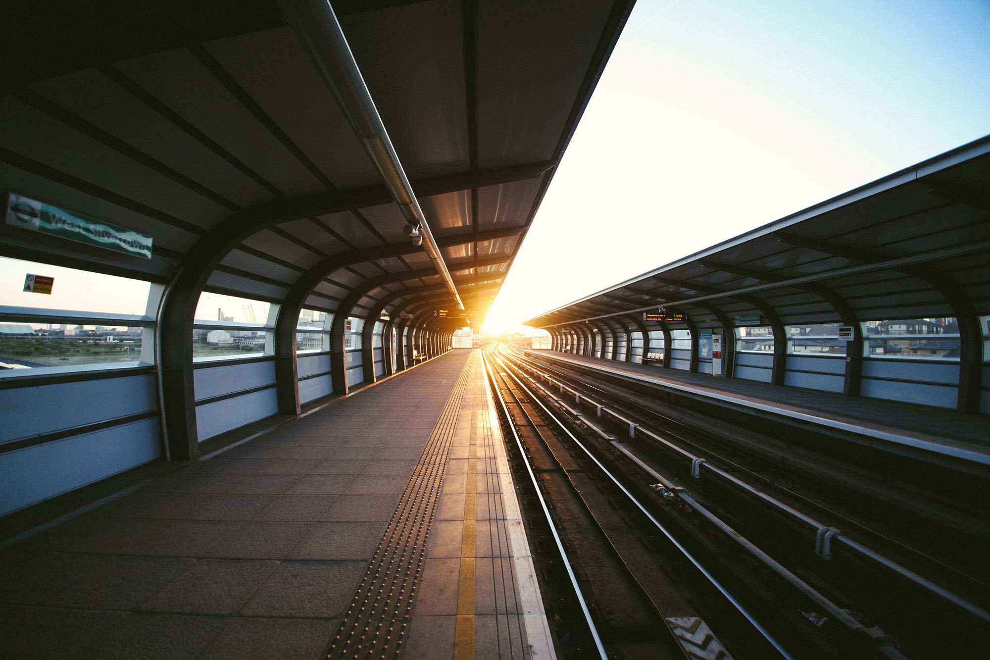 An open air railway station with the tracks heading off towards a sunset or sunrise on the horizon