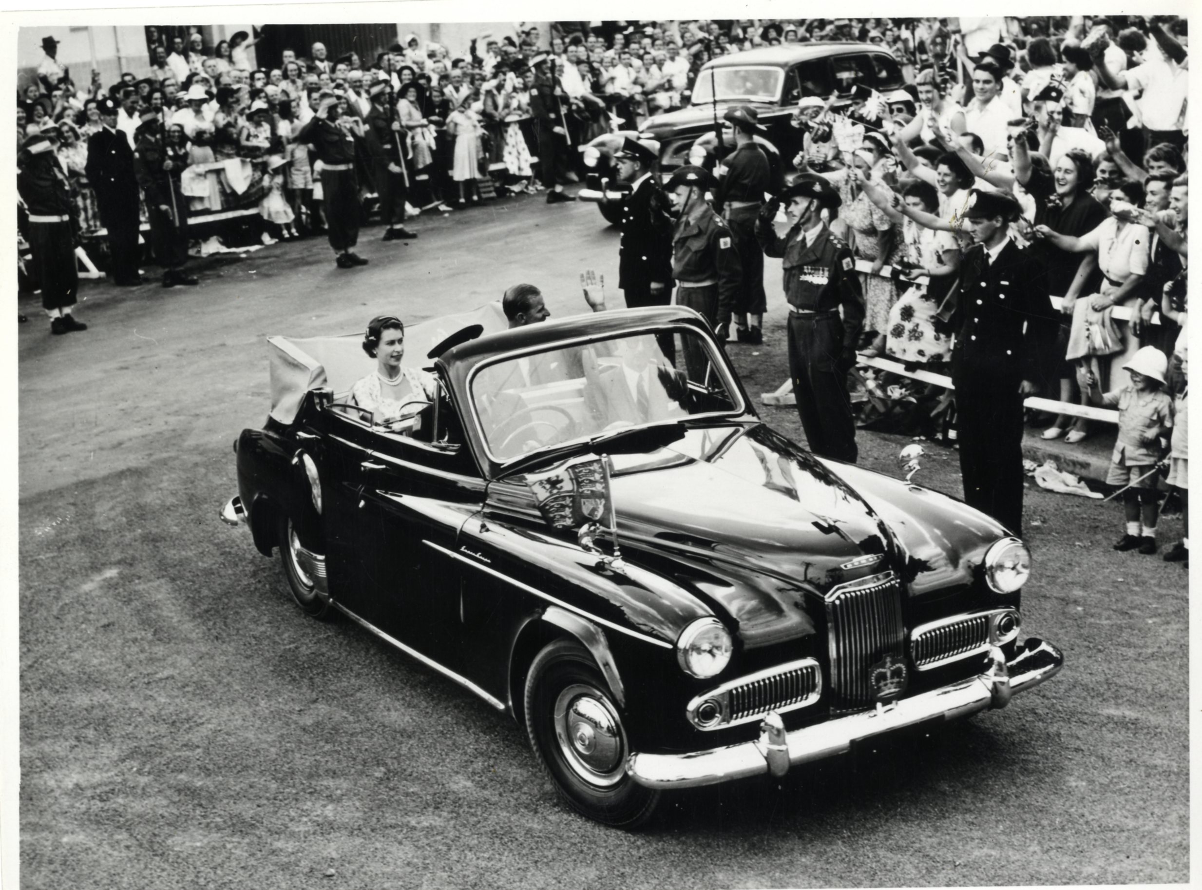 A black and white photograph of Queen Elizabeth II and the Duke of Edinburgh driving past crowds of people in an open-top Humber car