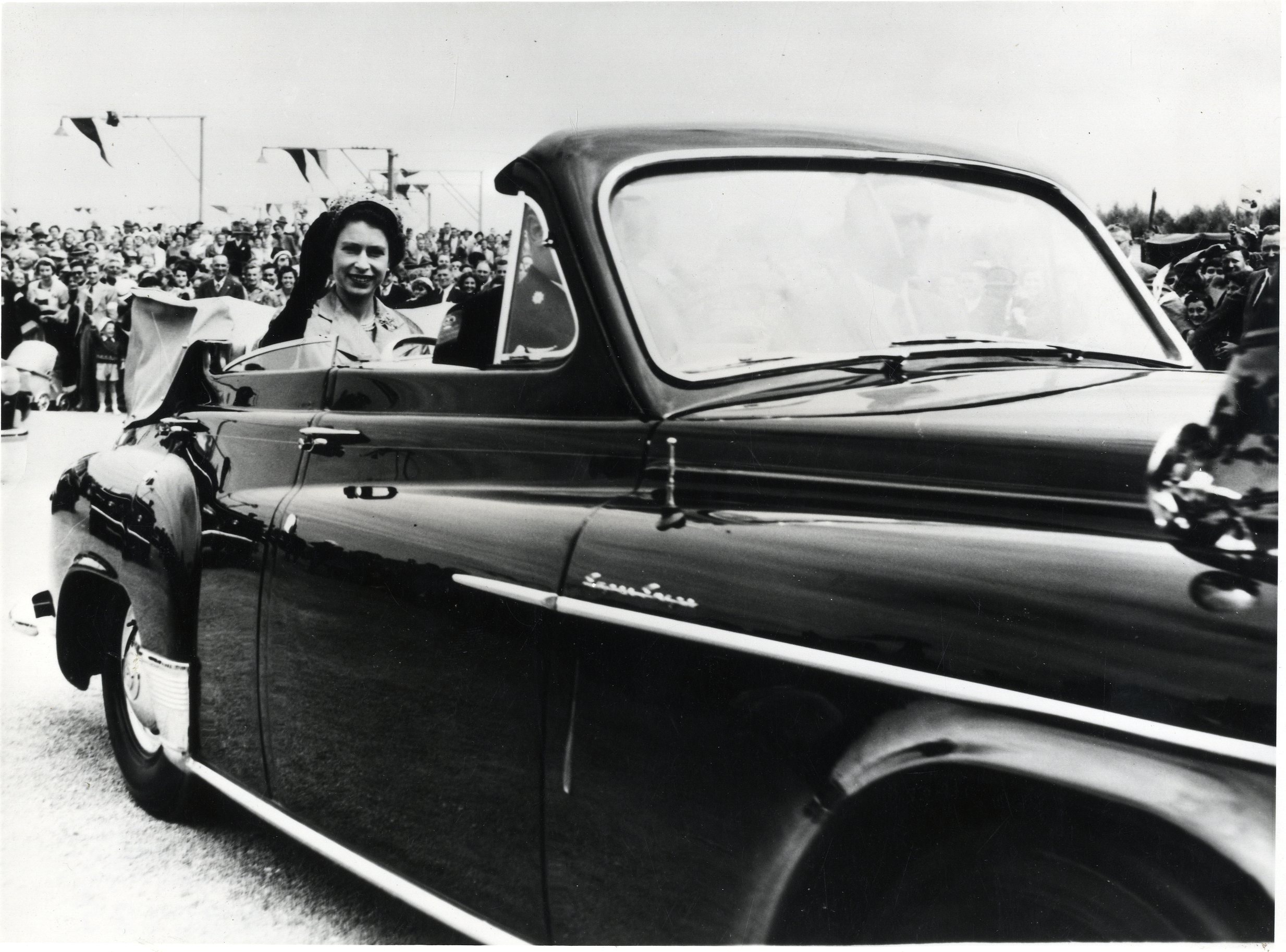 A black and white photograph of HM Queen Elizabeth II in an open top car, smiling and waving towards the camera. A crowd of people have gathered to watch, and there is bunting hanging from street lamps in the background.