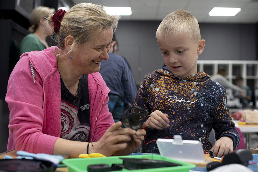 A photo of a smiling woman and a boy sitting at a table covered in tools and electronic parts.