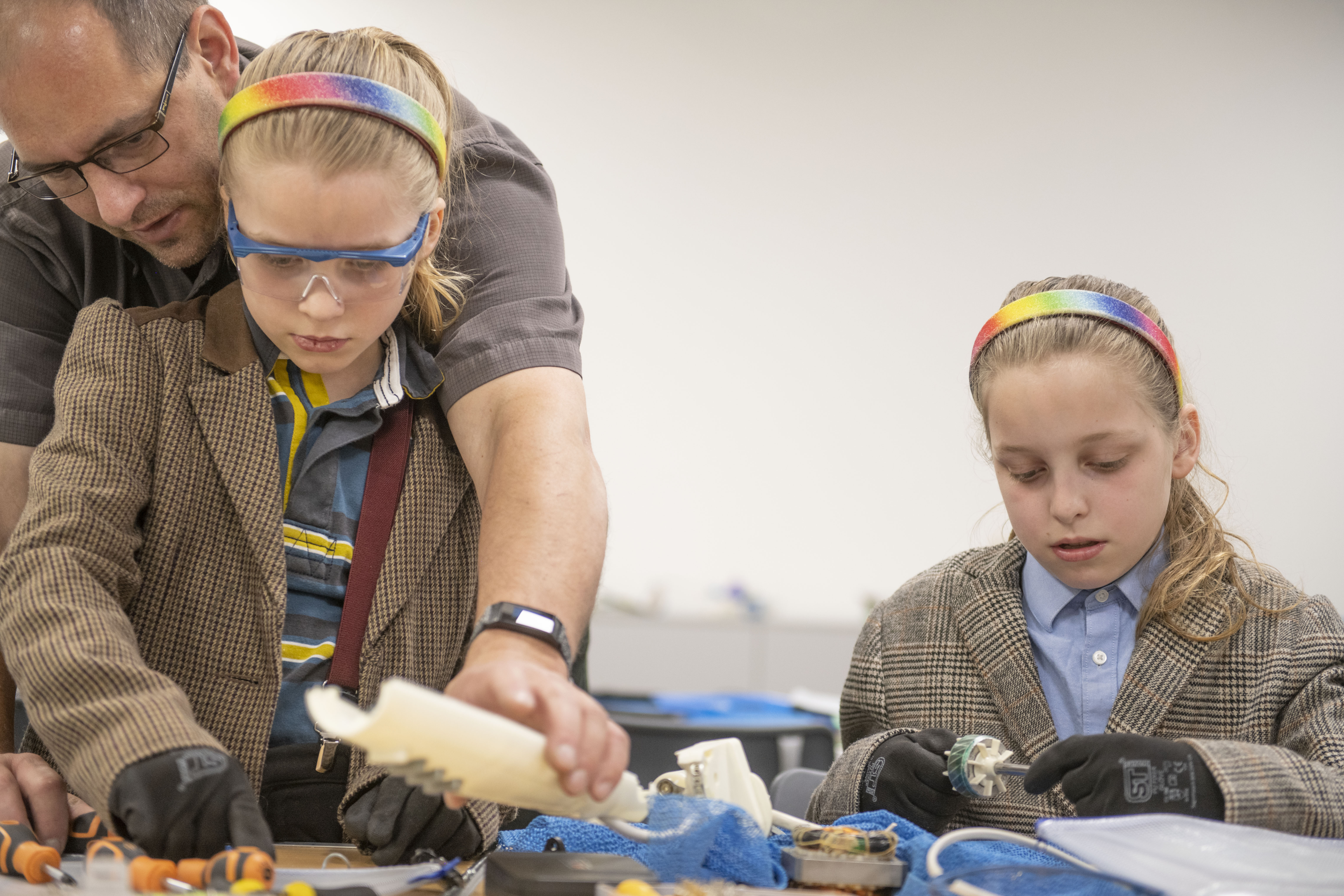 Two girls with ponytails, blazers and and matching rainbow coloured headbands are working with tools at a table. Both are wearing protective gloves and the girl on the left is wearing safety goggles. Behind her, a man helps her with some of the objects on the table.